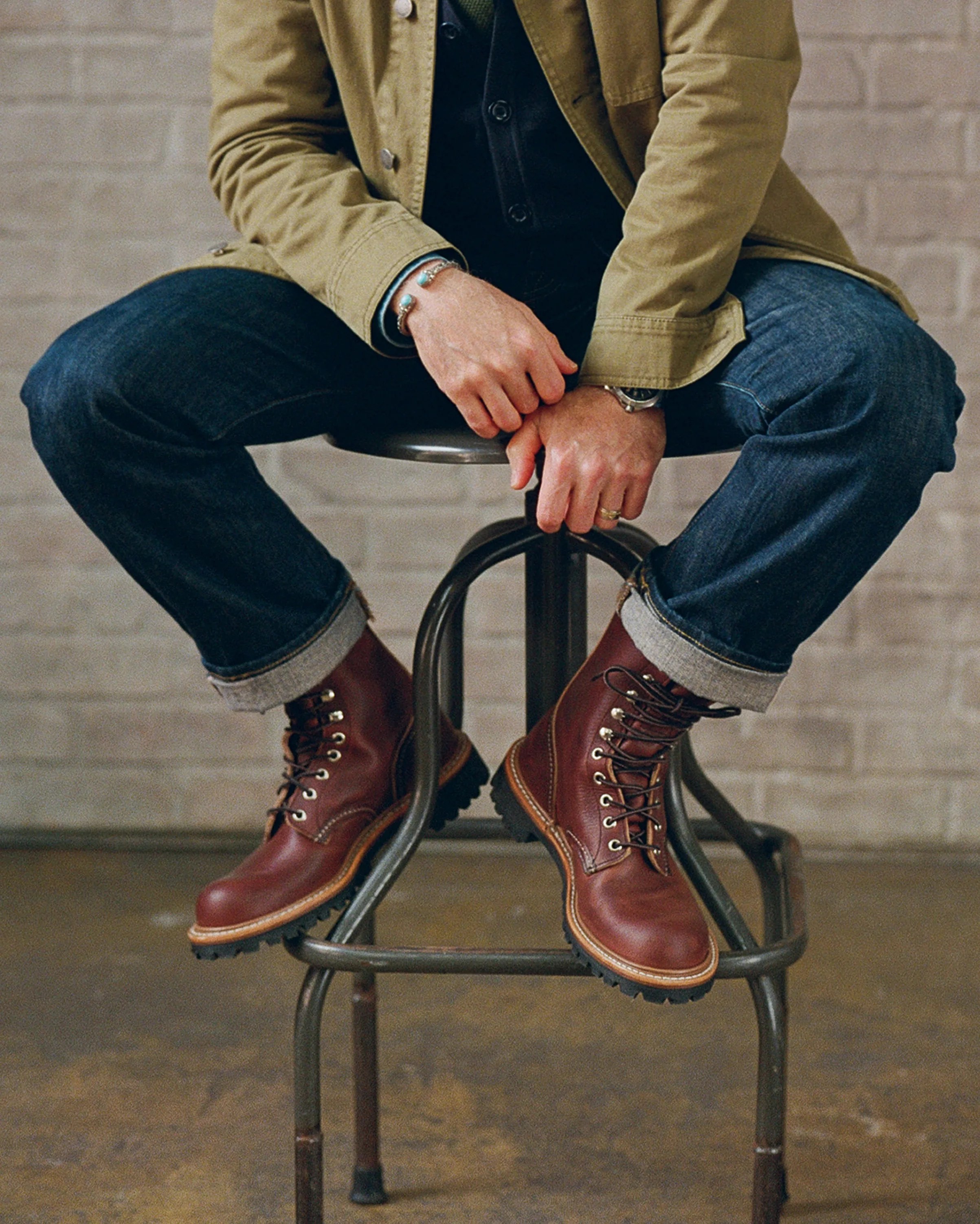 Person wearing rolled-up dark blue jeans and brown leather lace-up boots sitting on a metal stool.
