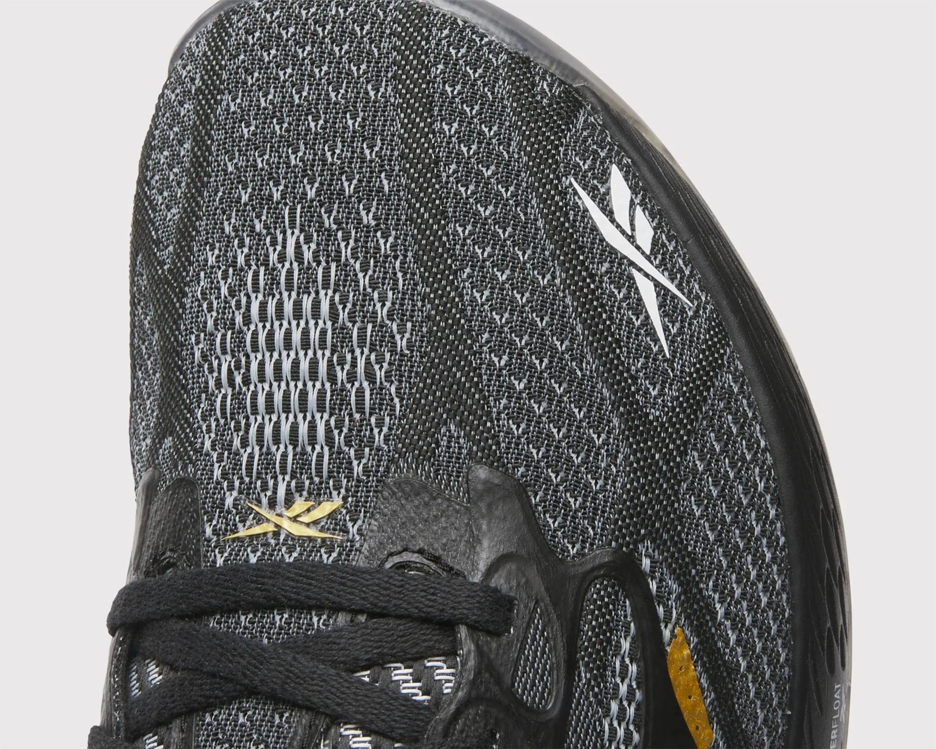 Close-up of black and gray knit sneaker upper with black laces, white and gold Reebok logos, and black sole edge.