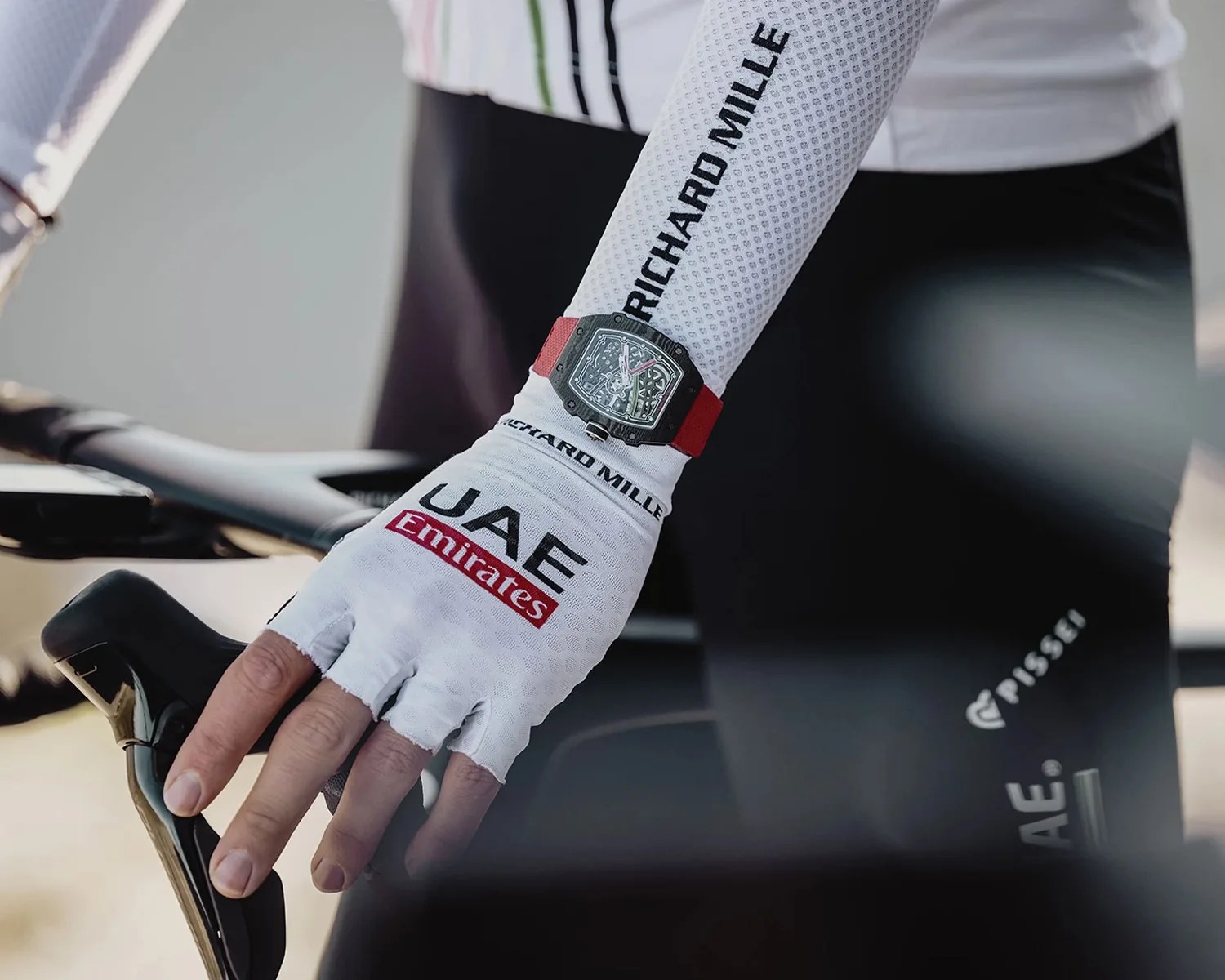 Skeleton dial wristwatch with a red strap worn over a white cycling glove on a hand gripping a bicycle handlebar.