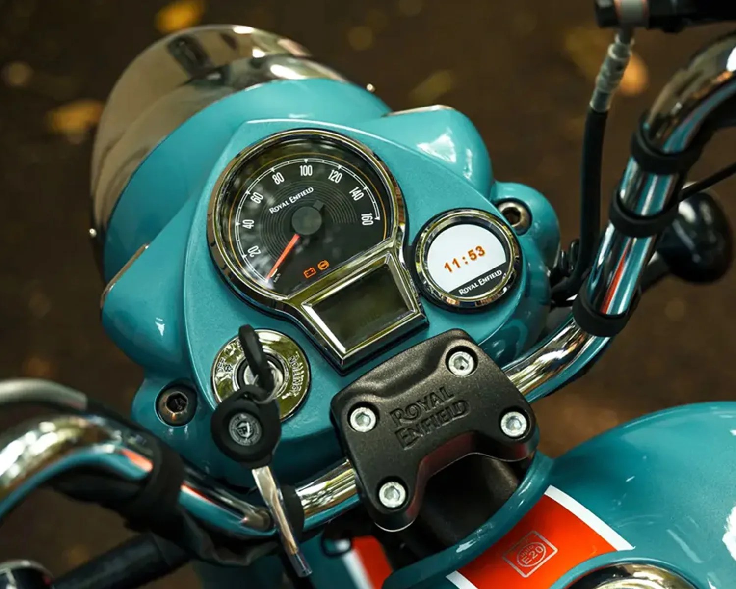 Close-up of a teal Royal Enfield motorcycle dashboard showing speedometer, digital clock, ignition key, and chrome handlebars.