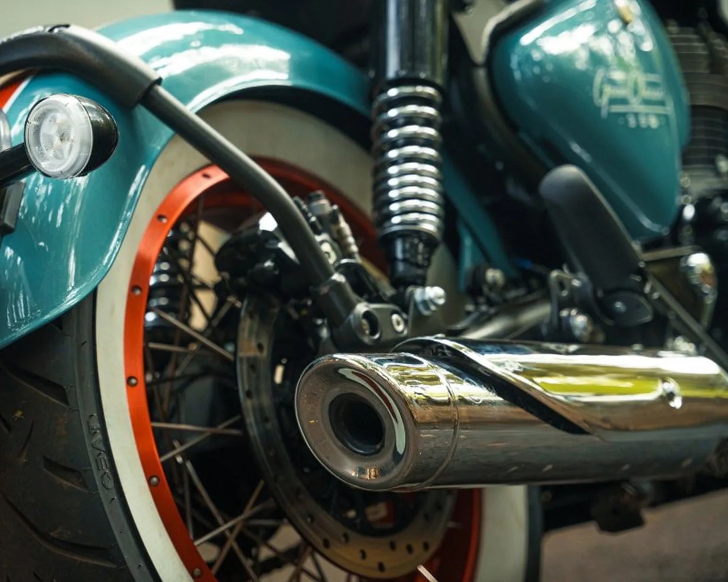 Close-up of a teal motorcycle rear wheel with whitewall tire, orange rim, chrome exhaust pipe, and suspension spring.