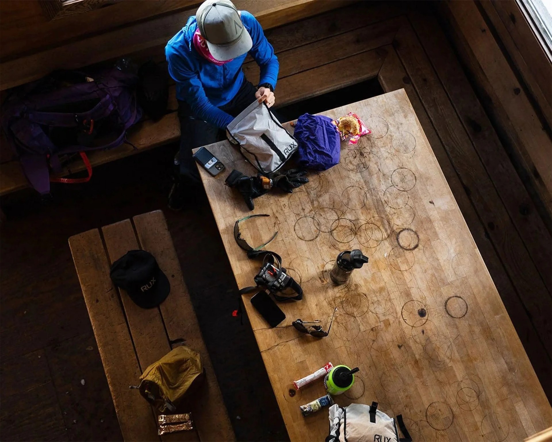 Person in blue jacket and gray cap sitting at a wooden table with outdoor gear and snacks scattered on it.