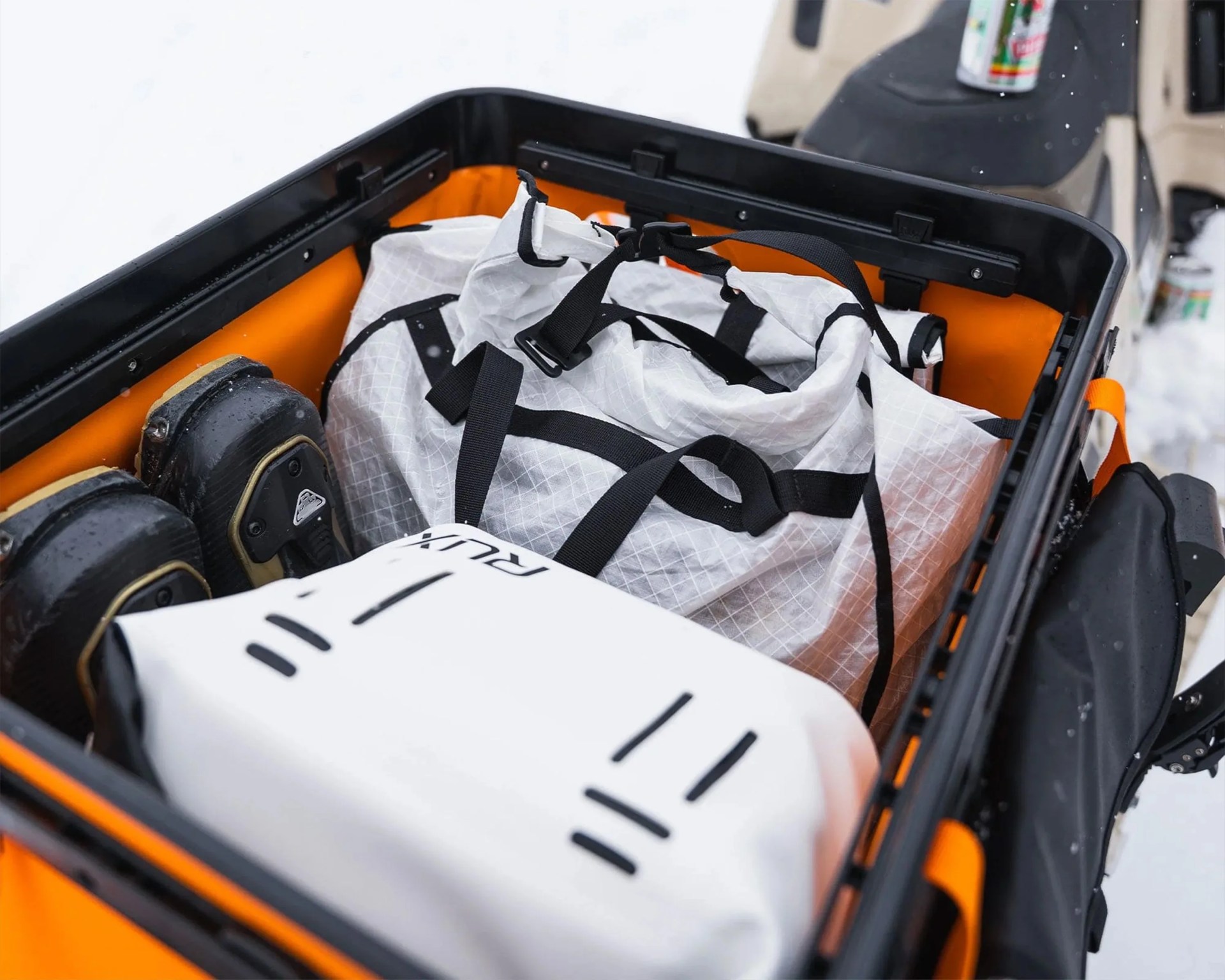 Orange and black cargo box containing a white duffel bag with black straps and a white bag with black markings, set in a snowy environment.