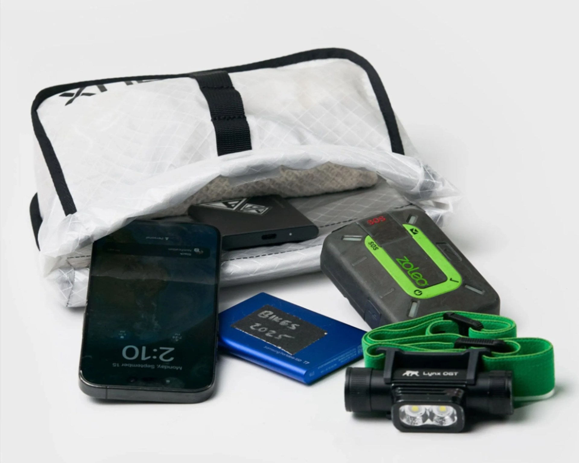 White zippered pouch with black trim containing a smartphone, blue battery pack labeled "Bikes 2025," green and black Zoleo SOS device, and black Lynx OGT headlamp with green strap.
