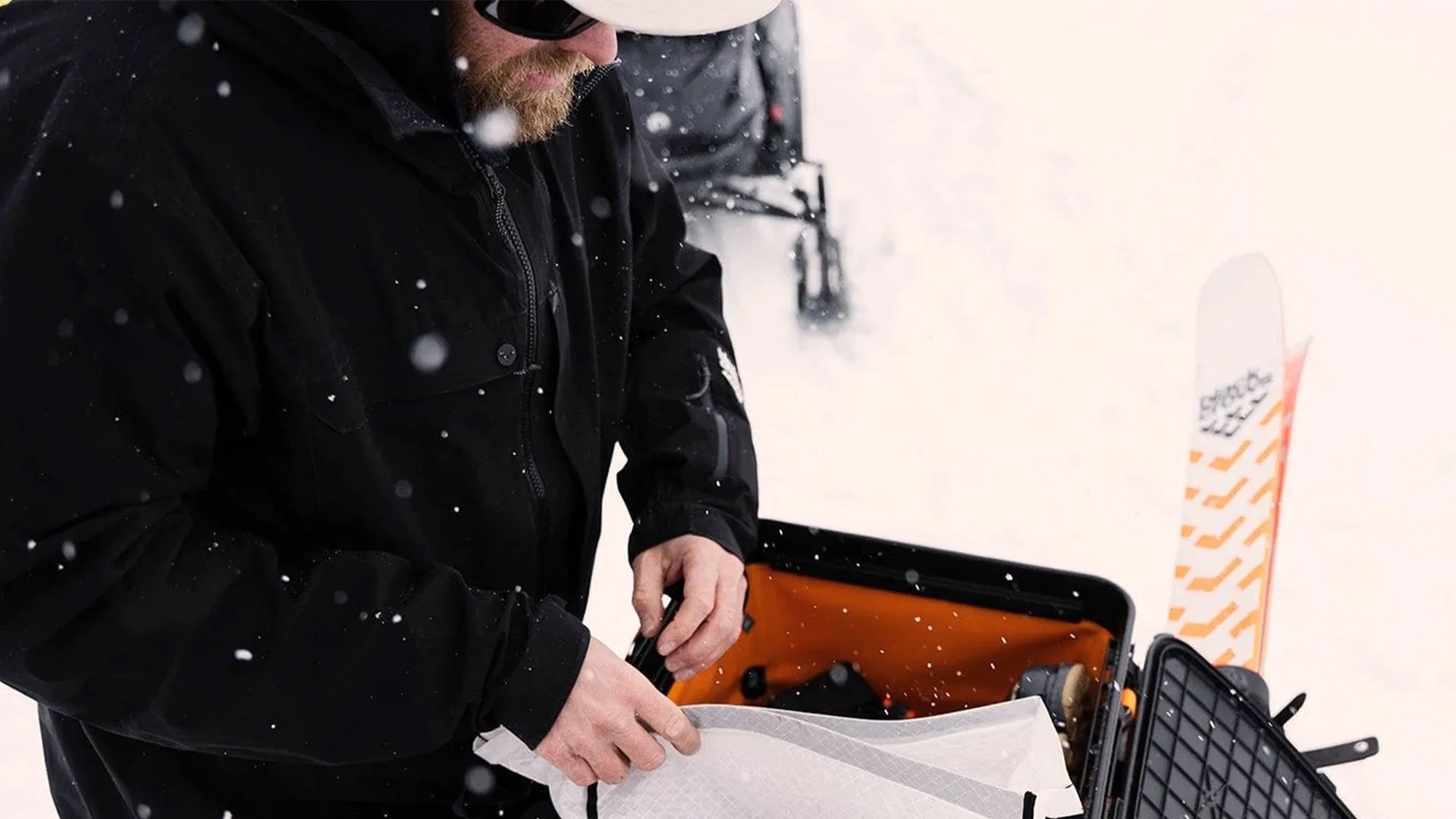 Person in a black jacket handling a white item near an open black and orange suitcase in falling snow.