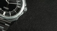 Close-up of a black dial automatic watch with silver hour markers and a stainless steel bracelet on a textured black surface.