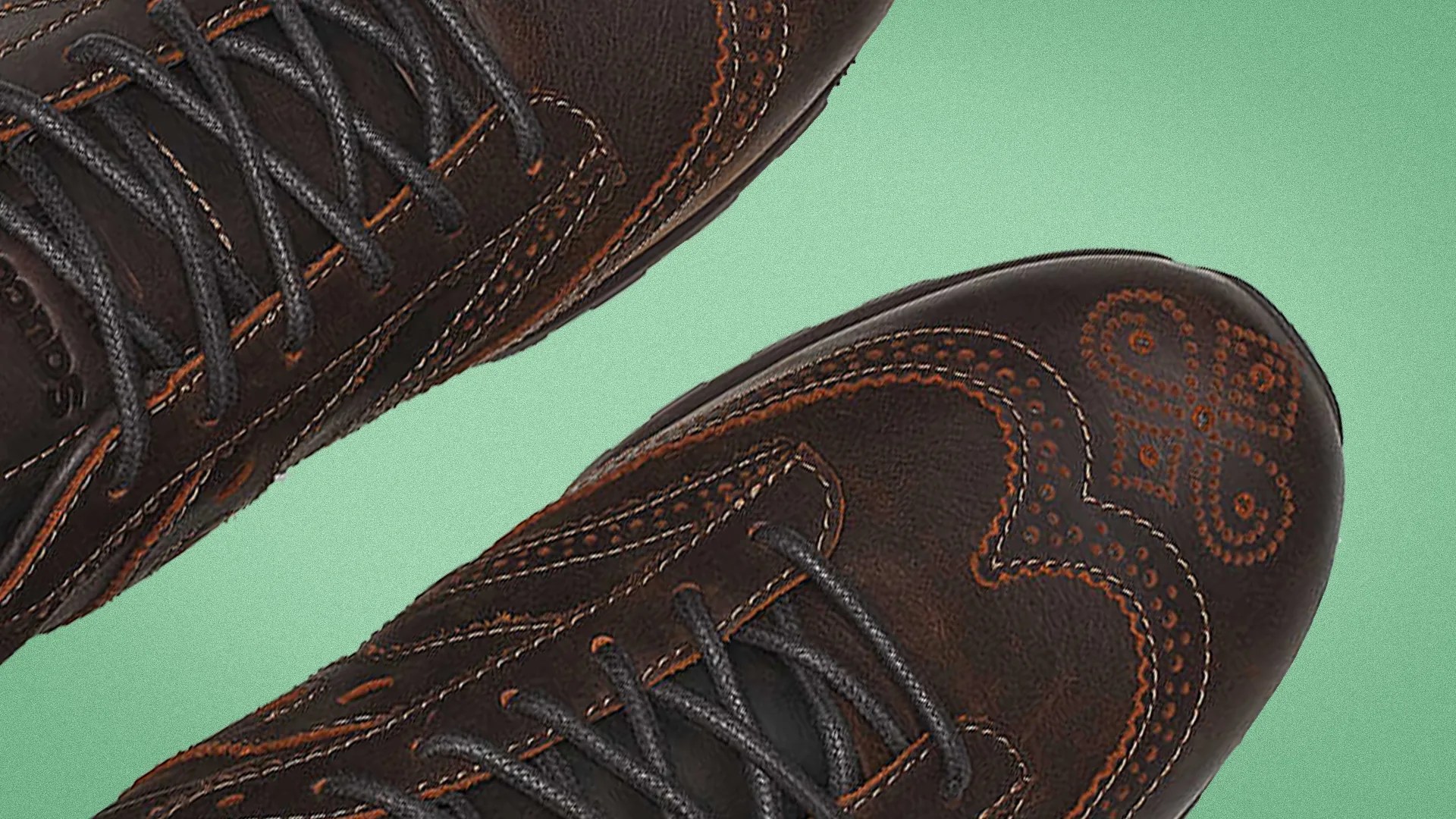 Close-up of dark brown leather shoes with orange decorative stitching and black laces on a green background.