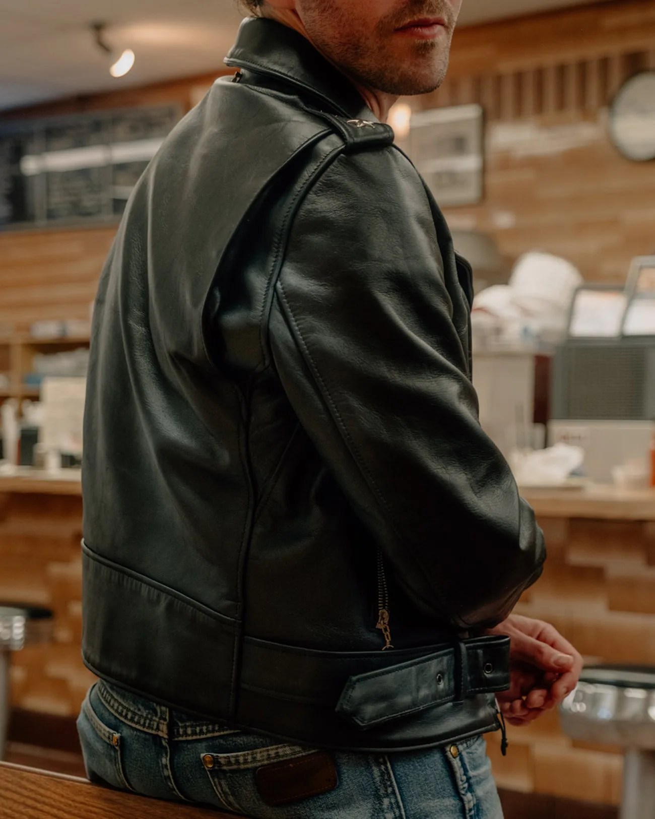 Black leather biker jacket with zipper and belt worn by a person sitting on a wooden bench.