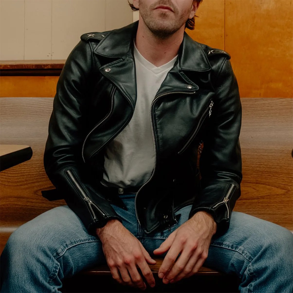 Black leather biker jacket with silver zippers worn over a white V-neck shirt and blue jeans.