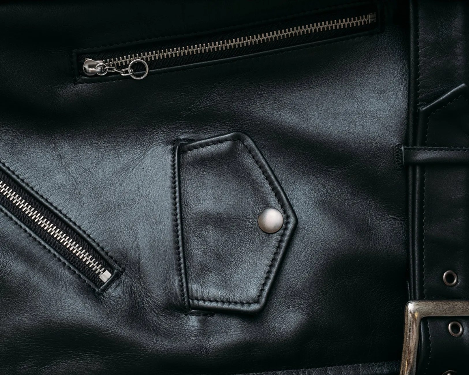Close-up of black leather jacket with silver zipper, snap button pocket, and belt buckle.