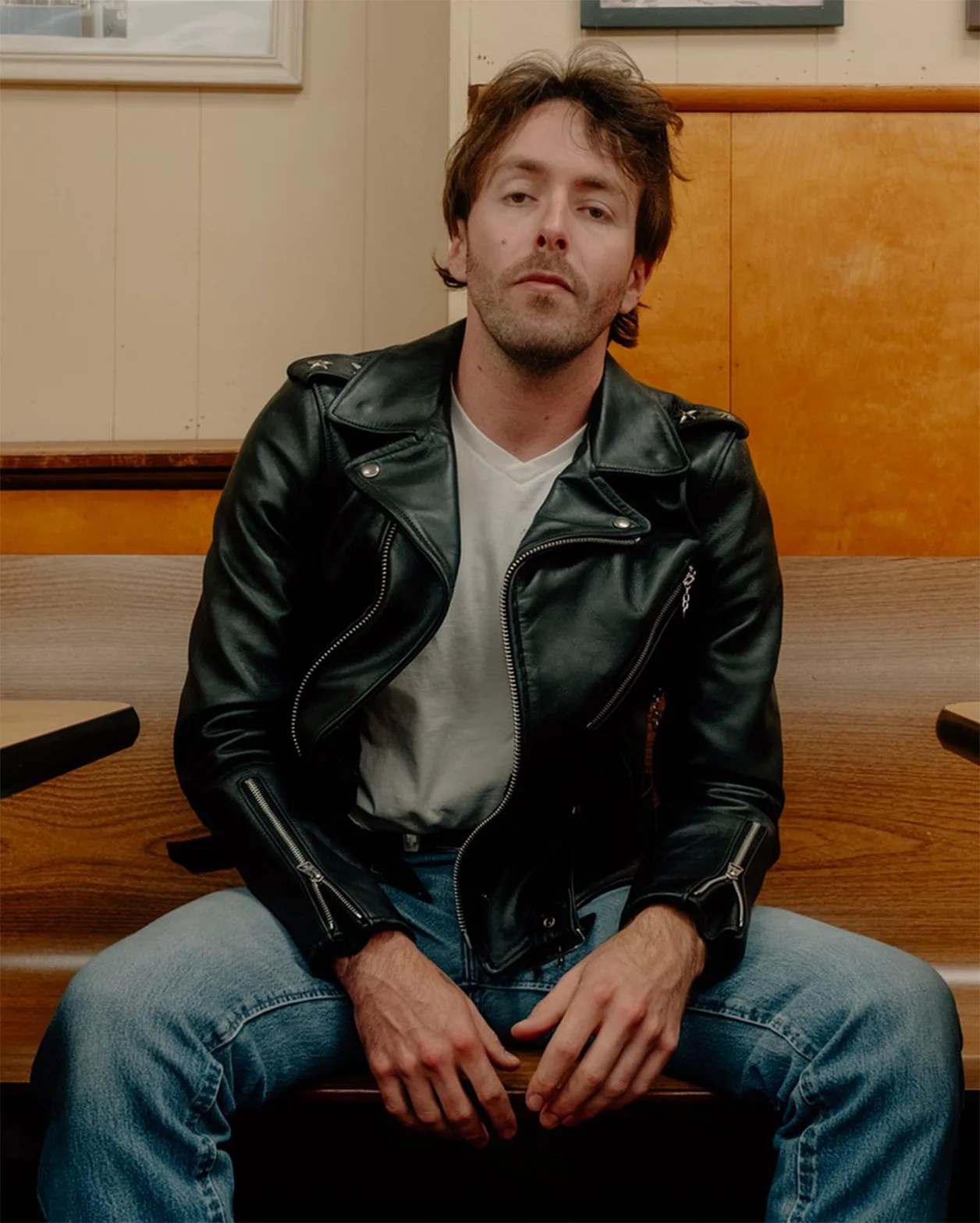 Man wearing a black leather jacket over a white t-shirt and blue jeans sitting on a wooden bench.
