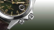 Close-up of a silver Seiko watch case with a green dial, gold numerals, and two textured crowns on a brown leather strap.