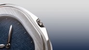 Close-up of a silver watch case with a textured blue dial and silver hands against a gradient gray-blue background.