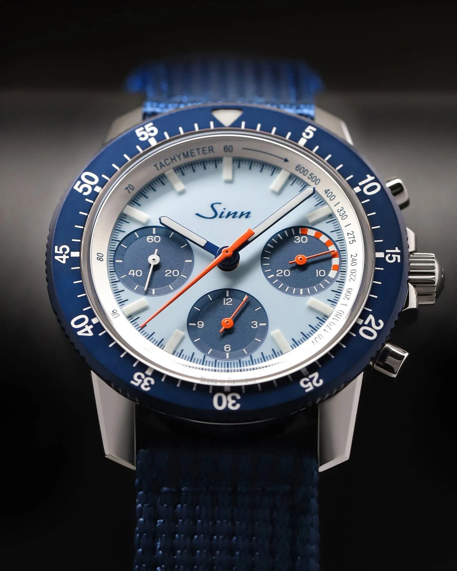 Blue and silver Sinn chronograph watch with orange hands and blue fabric strap on a dark background.