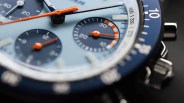 Close-up of a blue chronograph watch face with orange hands and a blue bezel with white markings.