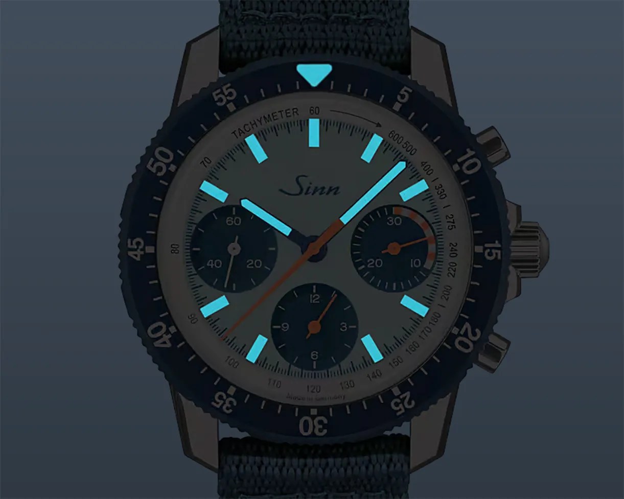 Sinn chronograph watch with black bezel, luminous blue hands and markers, and black fabric strap.