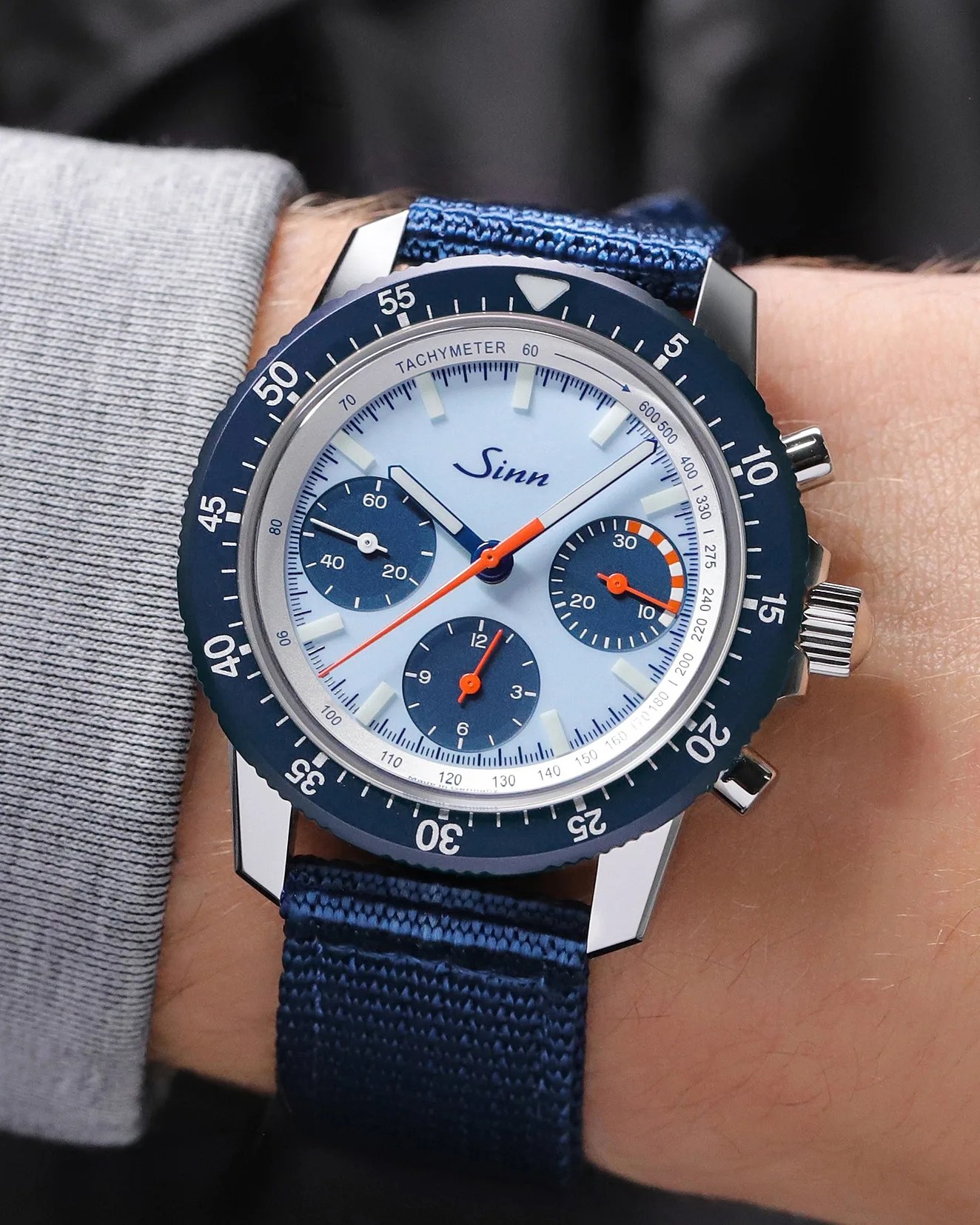 Blue Sinn chronograph watch with white dial, orange hands, and blue fabric strap worn on a wrist.