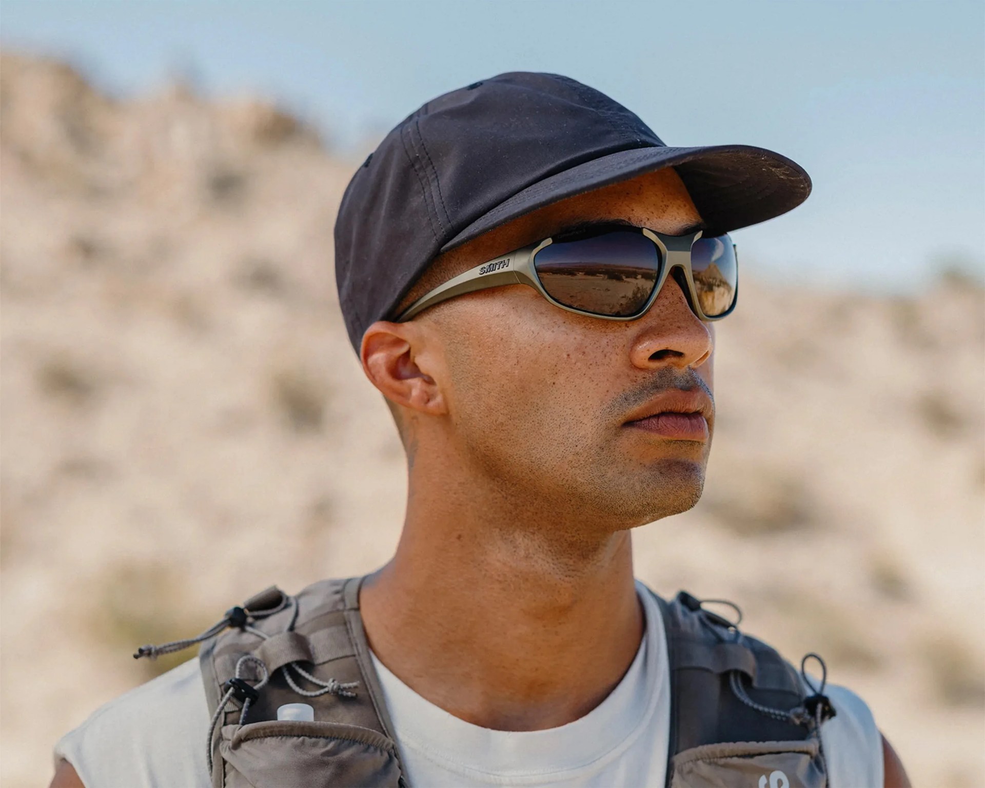 Man wearing matte black cap and gray Smith sunglasses with reflective lenses in a desert setting.