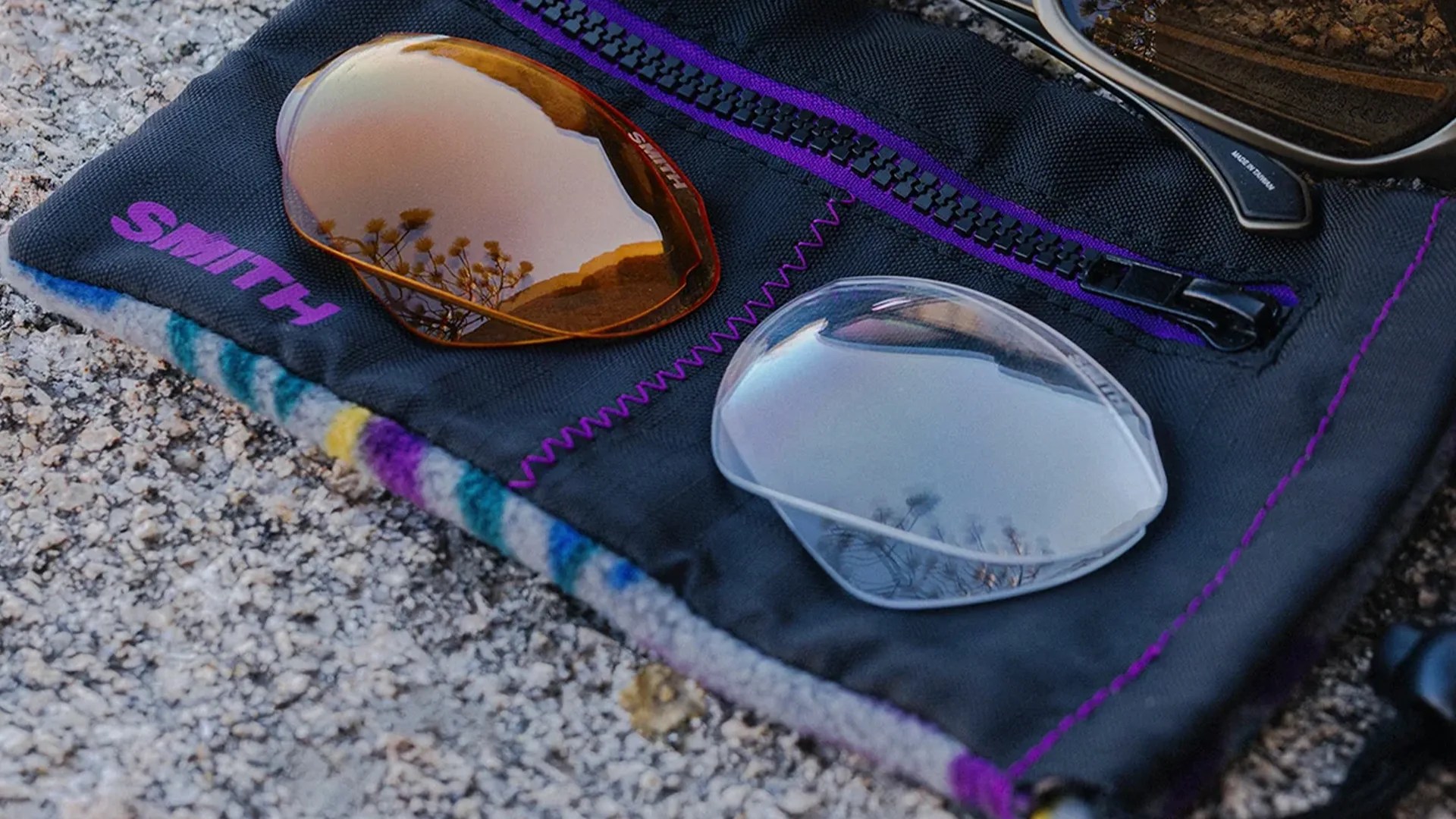 Two replacement lenses, one amber and one clear, resting on a black Smith branded pouch with purple stitching and zipper on a gravel surface.
