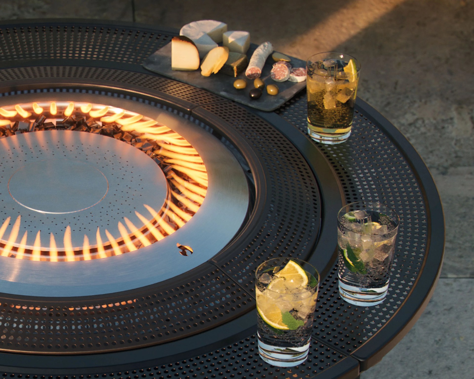 Round black metal fire pit table with perforated surface, three glasses with lemon and mint, and a cheese and charcuterie board.