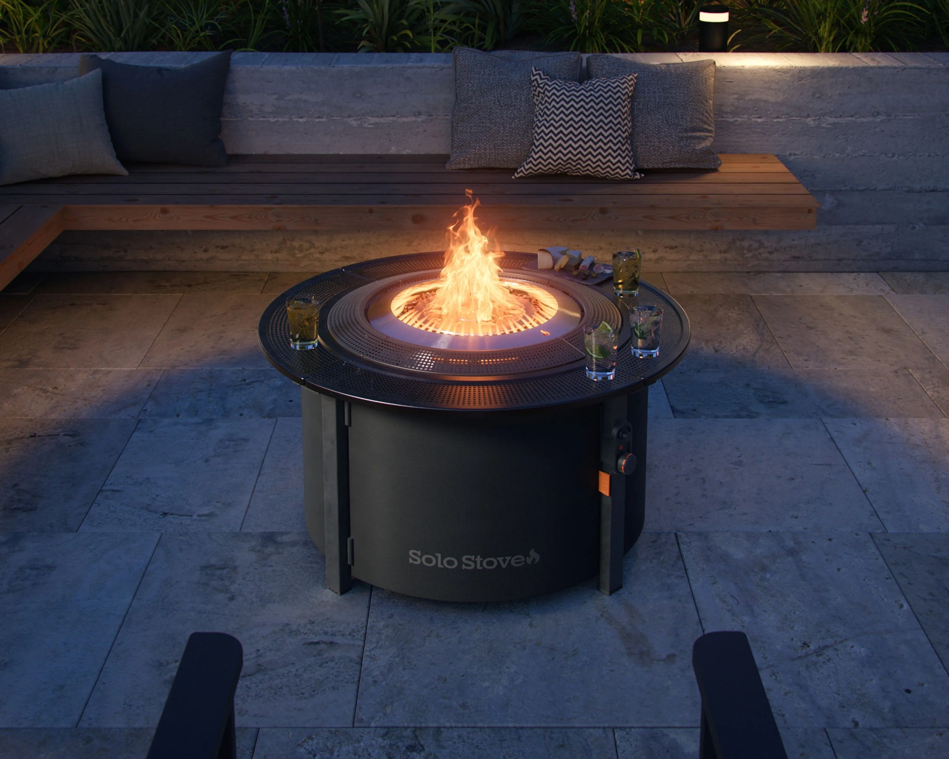 Round black Solo Stove fire pit with a flame burning, surrounded by four drinks and a wooden bench with cushions in the background.