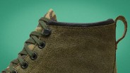 Close-up of an olive green suede boot with green laces and a camouflage fabric tongue against a green background.