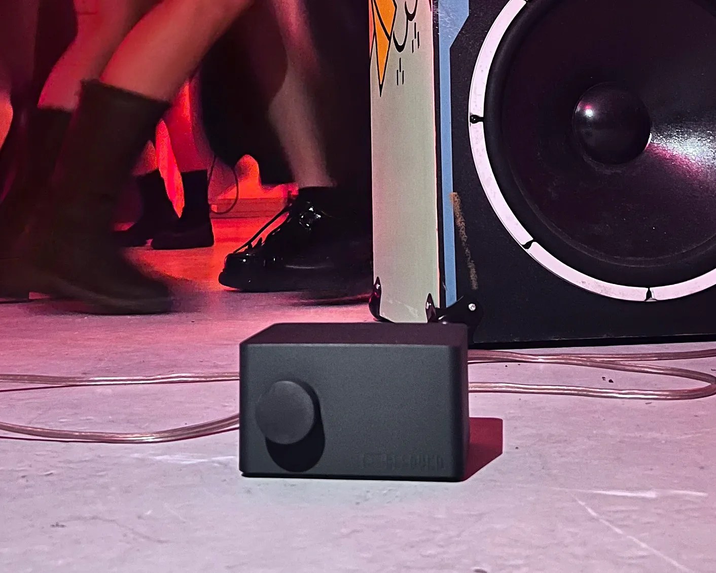 Black rectangular audio control box with a large knob on a floor near a speaker and people wearing boots.