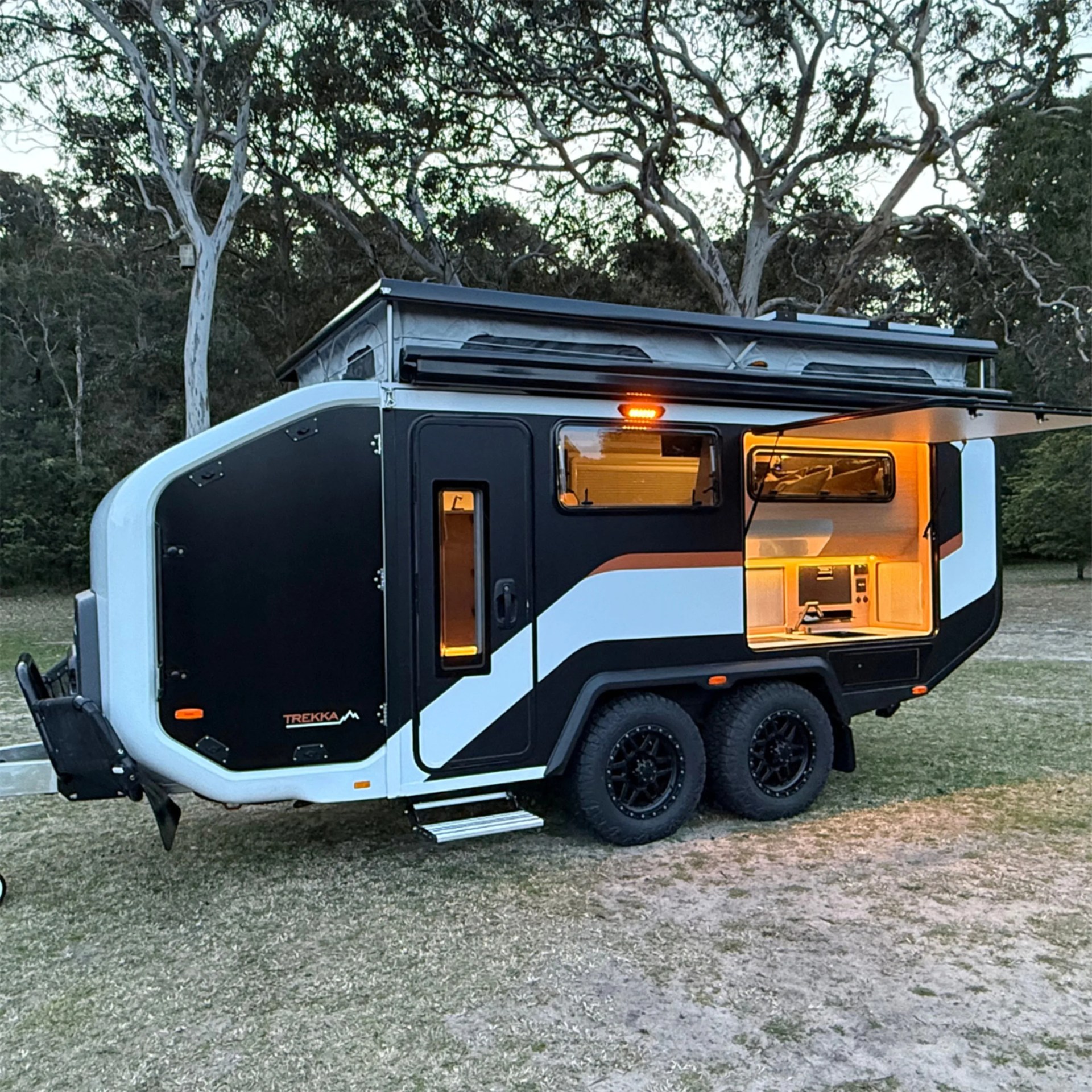 Black and white Trekka camper trailer with pop-up roof and illuminated outdoor kitchen in a wooded area.