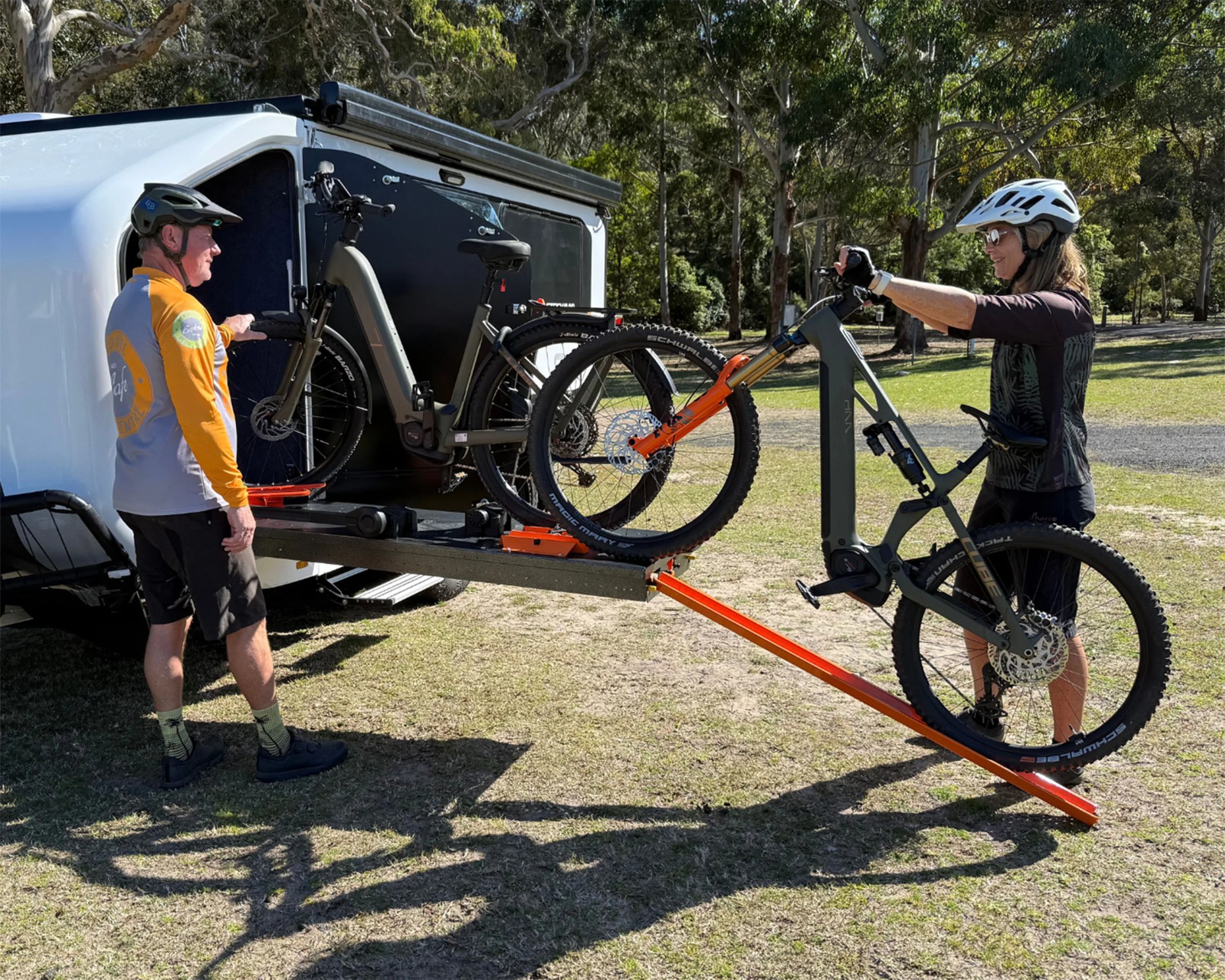 Two people loading electric mountain bikes onto a white trailer using an orange ramp in a grassy park.