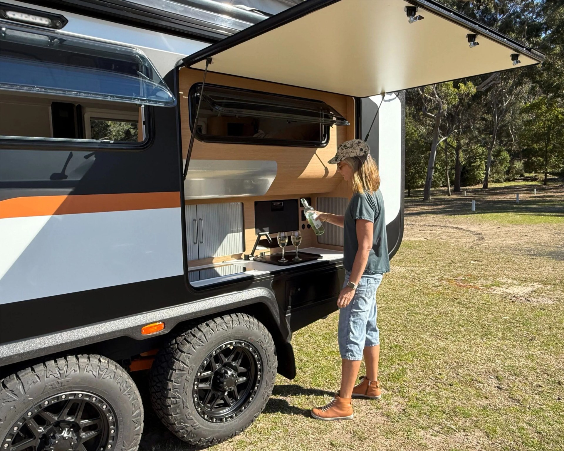 Person pouring wine into glasses from an outdoor kitchenette of a black and white camper trailer with off-road tires.