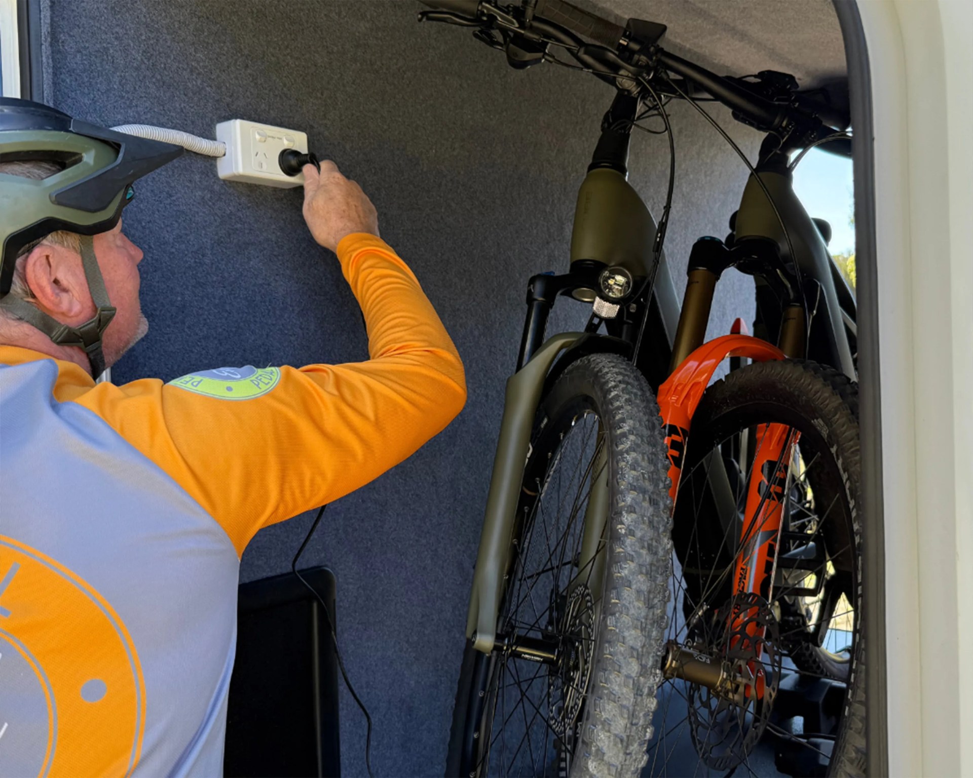 Person in an orange and gray cycling jersey plugging a cord into a wall socket inside a vehicle with two mountain bikes.