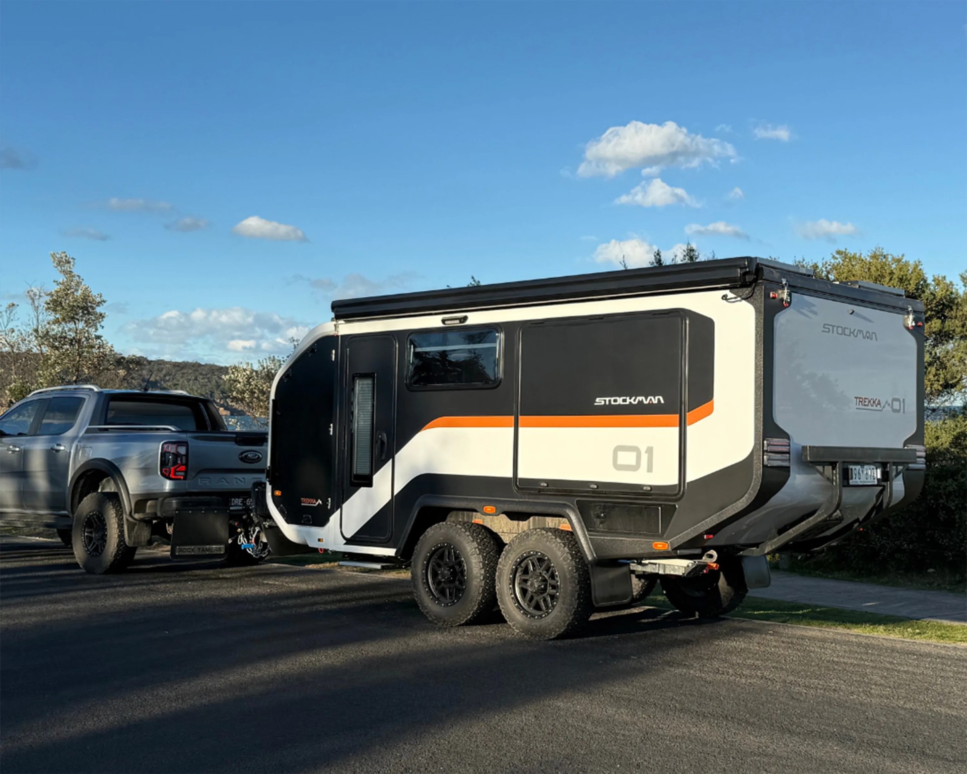 Black, white, and orange Stockman Trekka 01 camper trailer with dual rear wheels hitched to a silver pickup truck on a paved road.