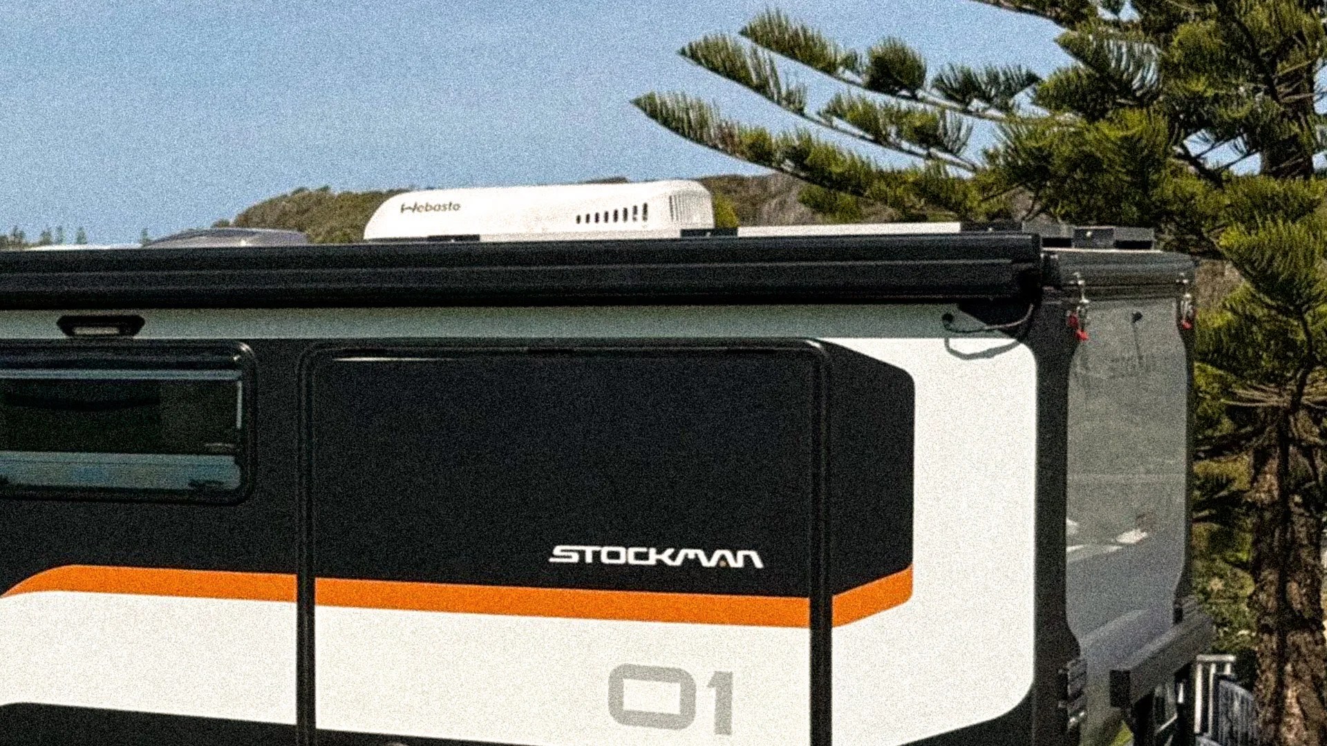 Side view of a black and white Stockman 01 camper trailer with an orange stripe and a Webasto roof unit.