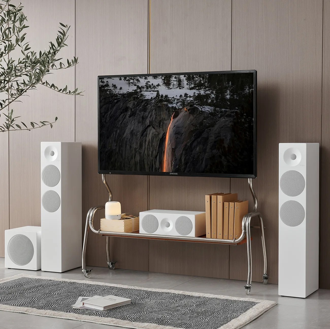 Modern living room setup with a flat-screen TV on a metal stand, white speakers, a subwoofer, books, and a small lamp on a wooden shelf.