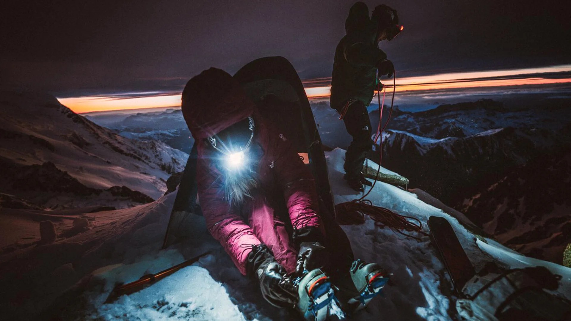 Two climbers on a snowy mountain at dusk, one sitting with a headlamp on and adjusting boots, the other standing with climbing rope.