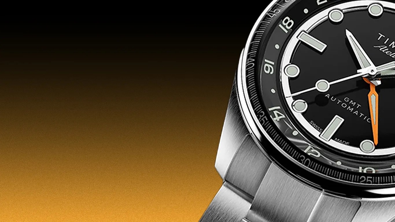 Close-up of a stainless steel wristwatch with a black dial, white markers, and an orange GMT hand.