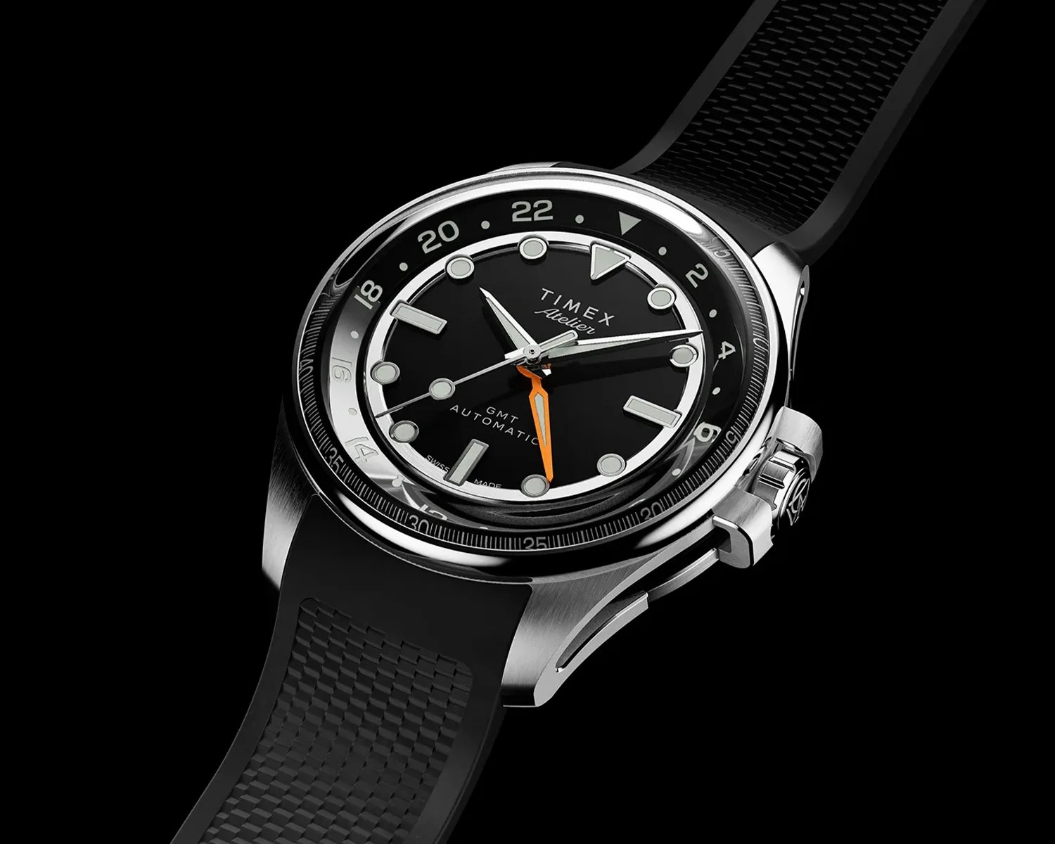 Stainless steel Timex Atelier GMT automatic watch with black dial, orange GMT hand, and black textured rubber strap.