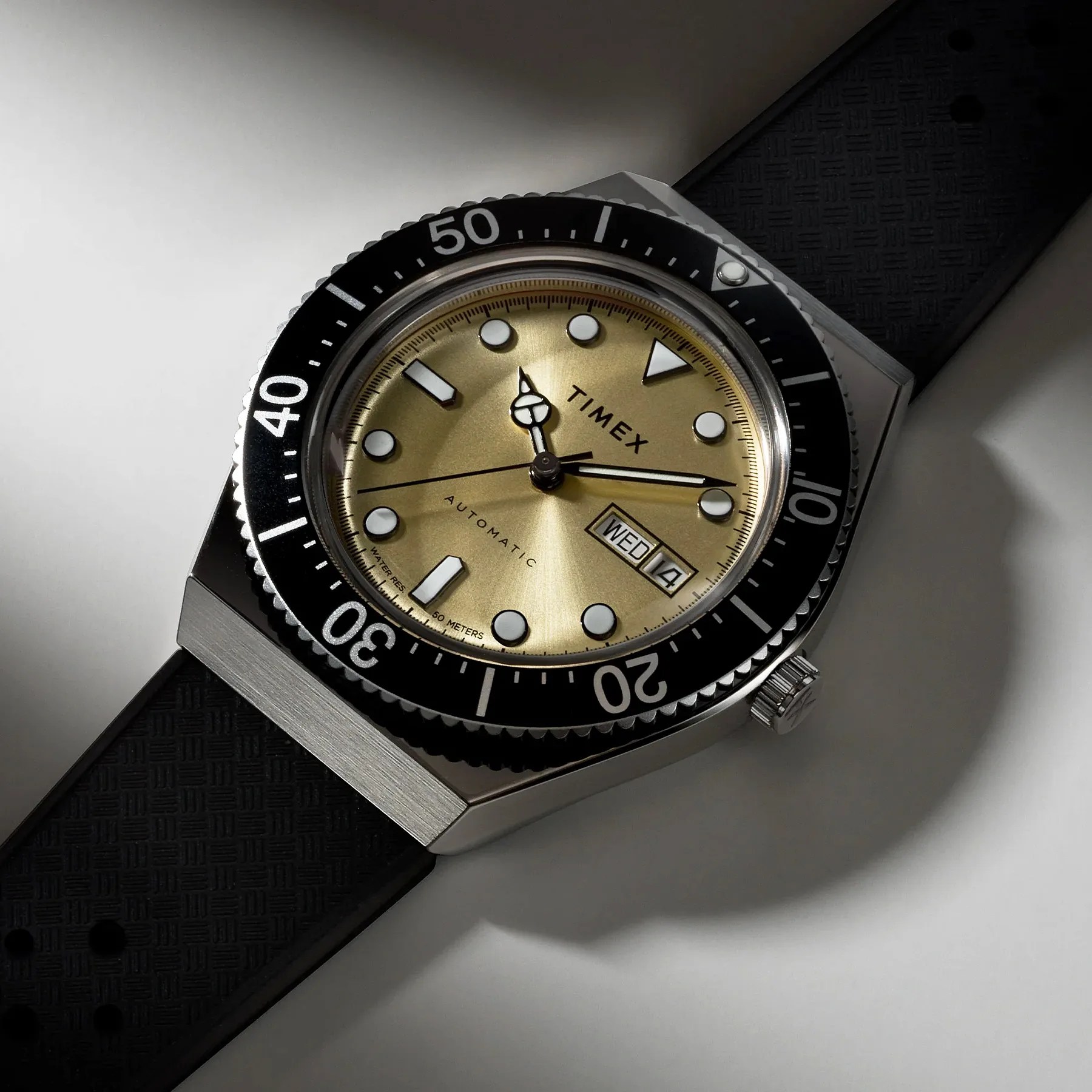 Timex watch with gold dial, black bezel, and black textured rubber strap on a gray surface.