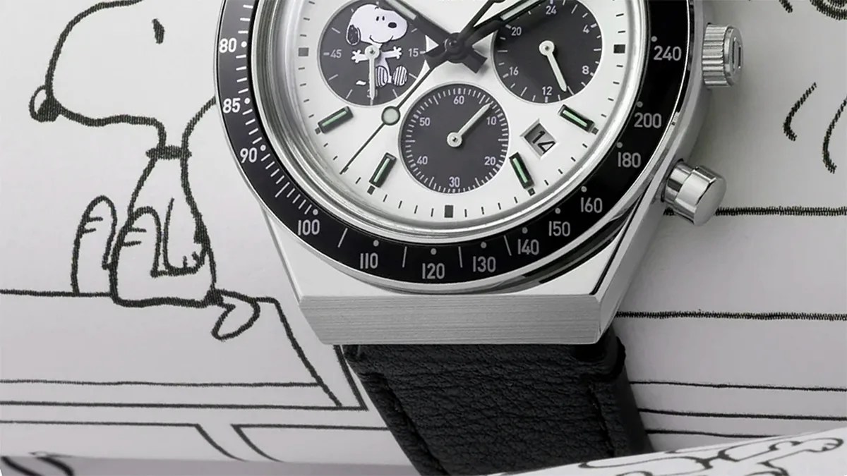 Close-up of a silver chronograph watch with black bezel, white dial featuring Snoopy illustration, and black leather strap.