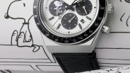 Close-up of a silver chronograph watch with black bezel, white dial featuring Snoopy illustration, and black leather strap.