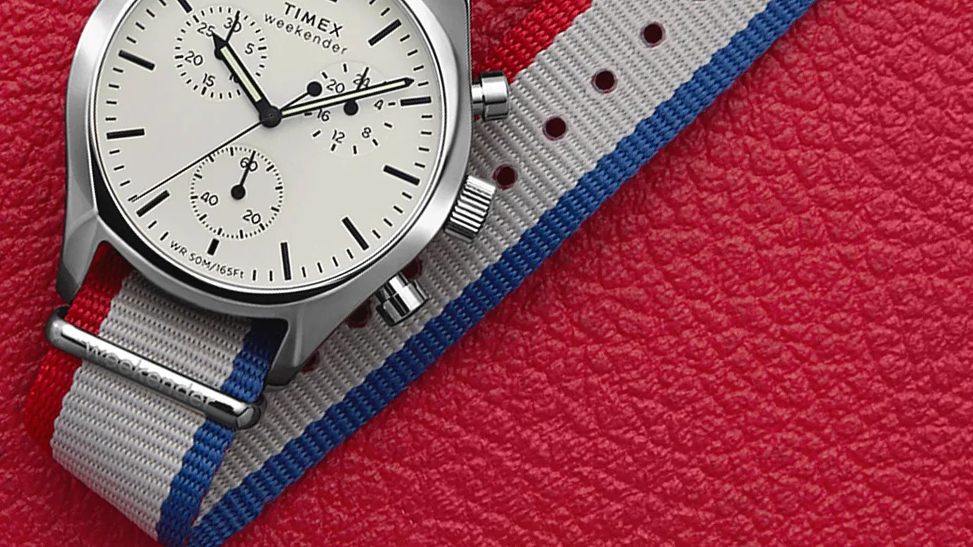 Silver Timex Weekender watch with white dial and red, white, and blue striped fabric strap on red textured surface.