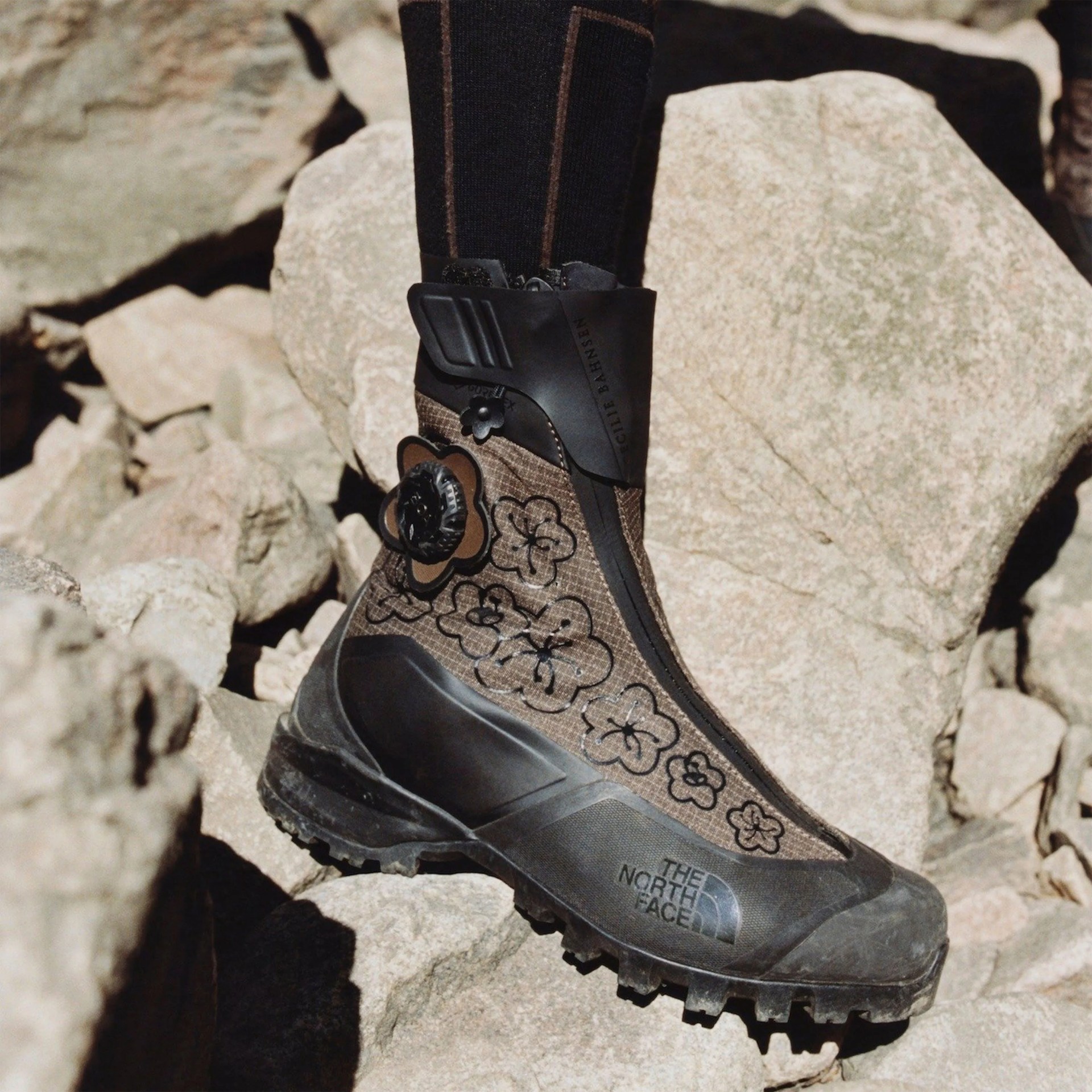 Brown and black rugged hiking boot with floral patterns and The North Face logo on rocky terrain.