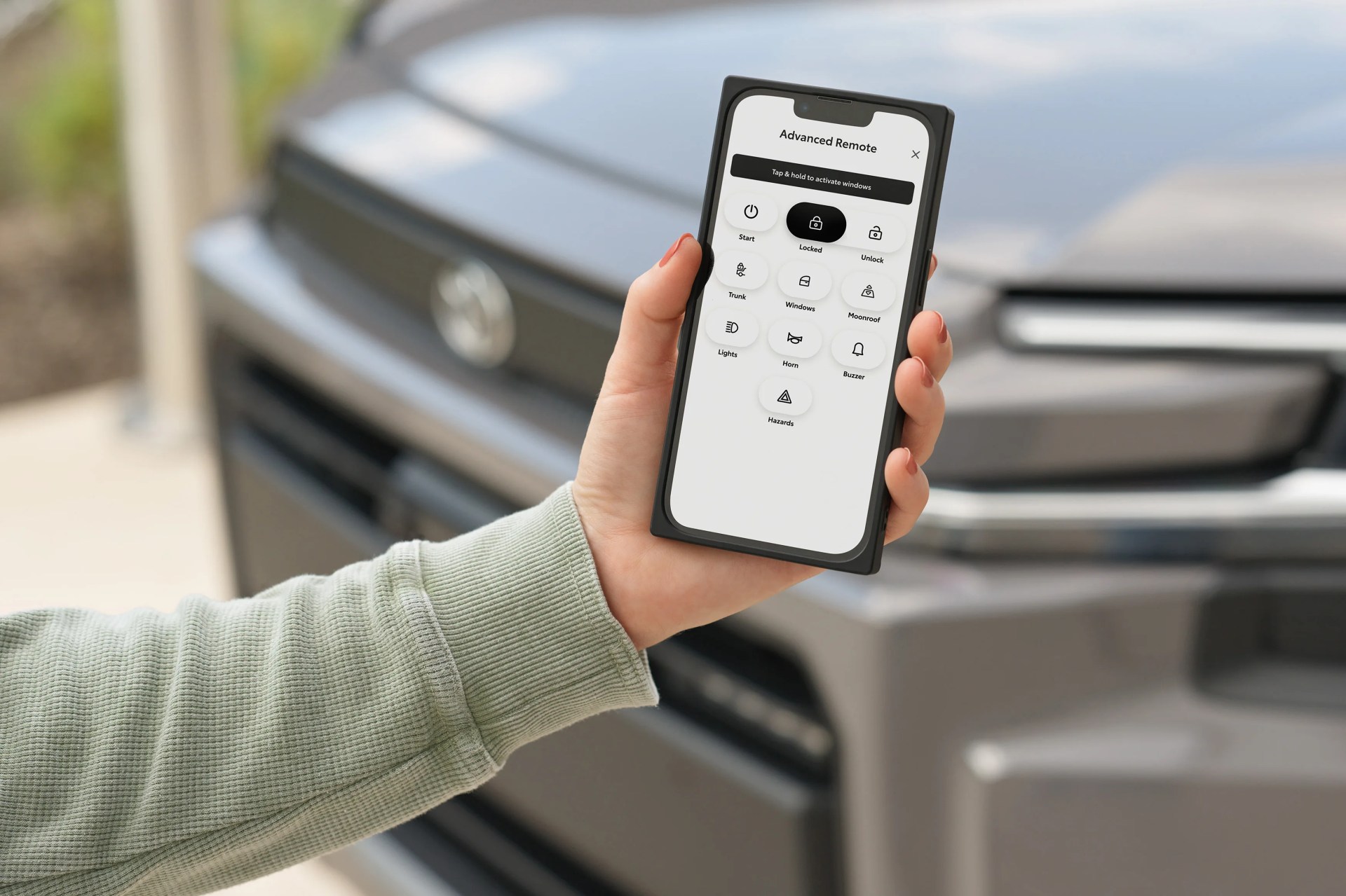 Hand holding a smartphone displaying an advanced remote control app in front of a gray Toyota vehicle.