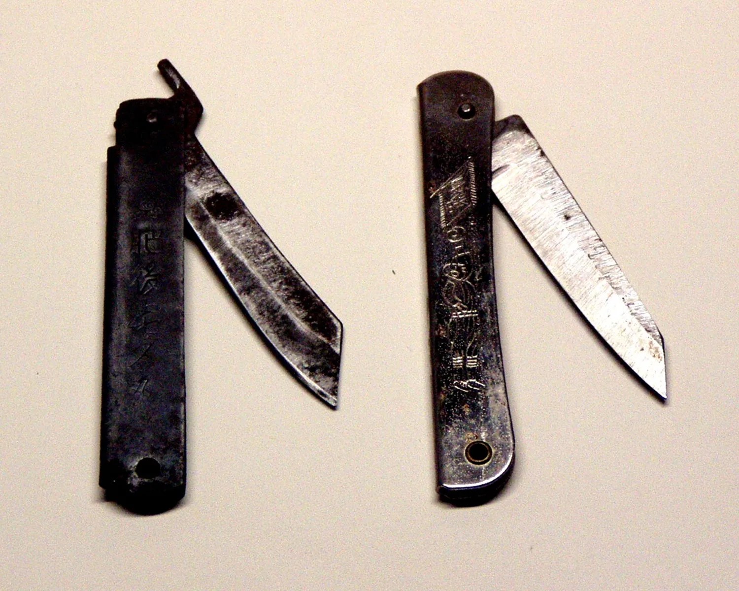 Two vintage black metal folding knives with partially open blades and engraved handles on a light surface.