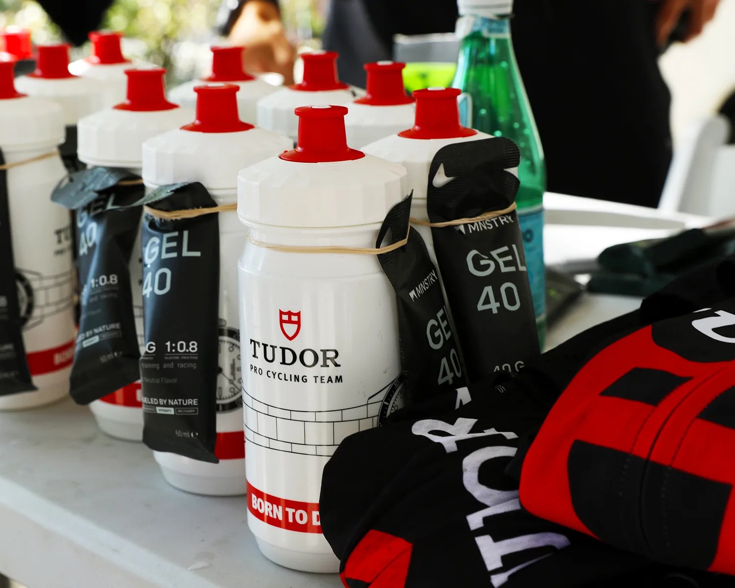 White Tudor Pro Cycling Team water bottles with red caps and attached black GEL 40 packets on a table.