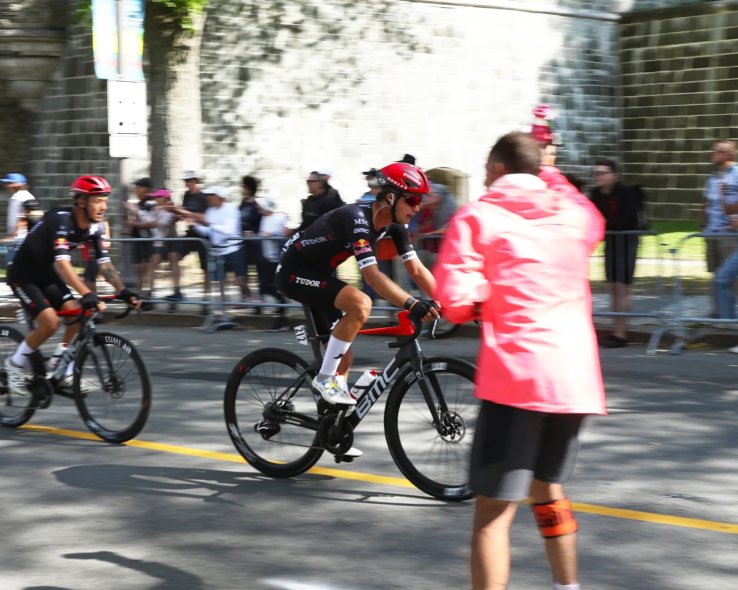Two cyclists in black and red racing gear riding BMC bikes on a road with spectators behind barriers.