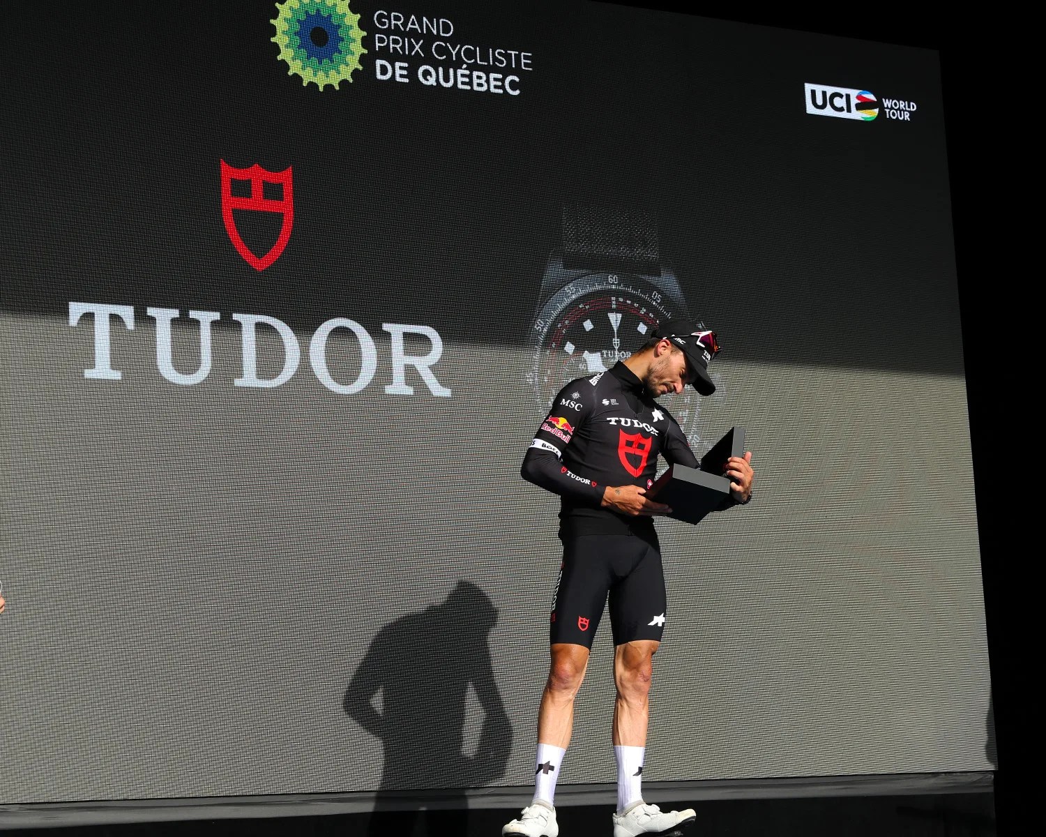 Cyclist in black Tudor-branded outfit holding an open black box on a stage with Tudor and Grand Prix Cycliste de Québec logos in the background.