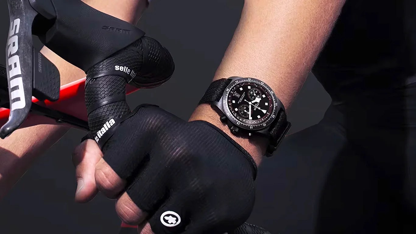 Black chronograph wristwatch with fabric strap worn on a wrist gripping a bicycle handlebar wrapped in black tape.