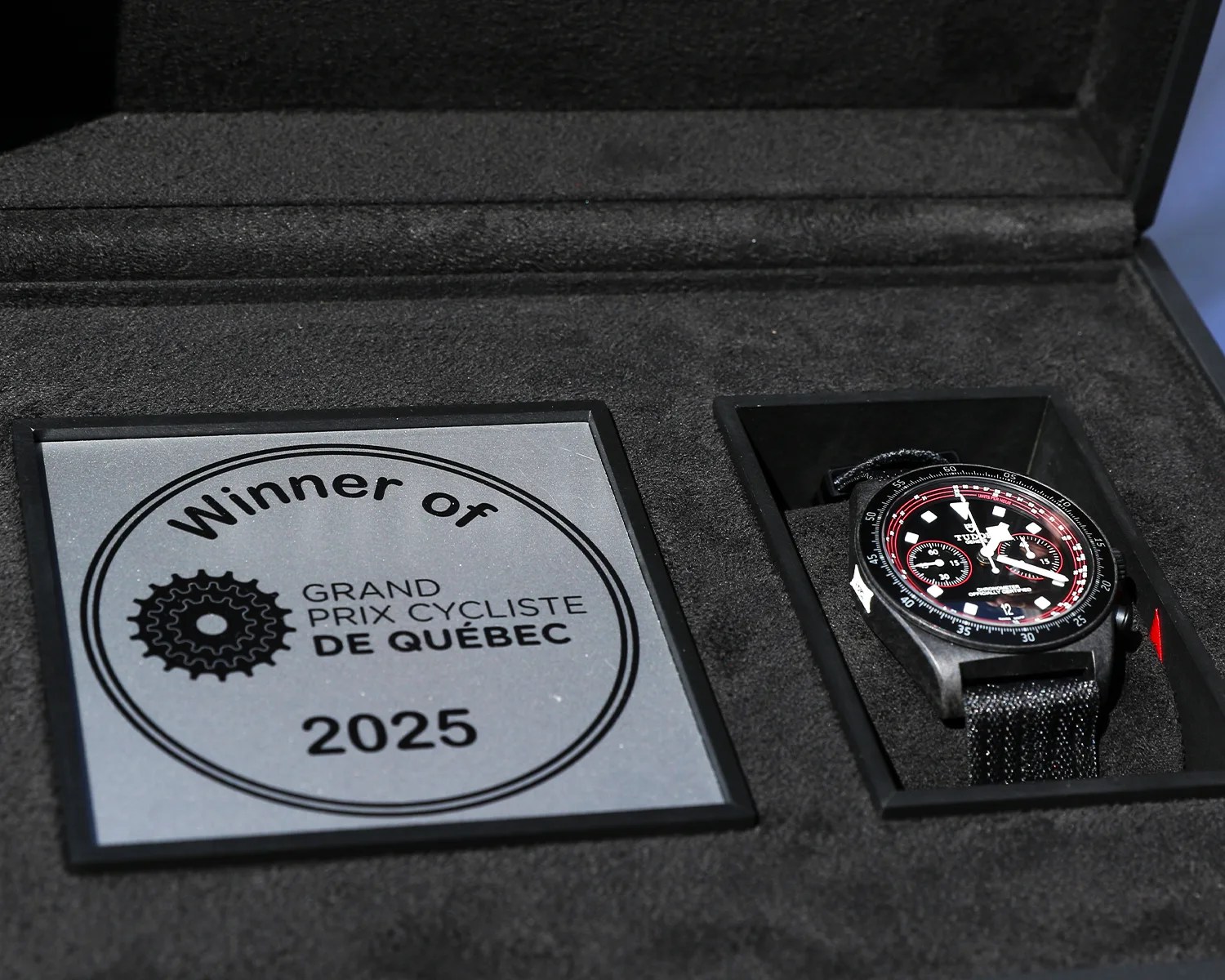 Black Tudor chronograph watch with red accents in a black presentation box next to a plaque reading "Winner of Grand Prix Cycliste de Québec 2025.