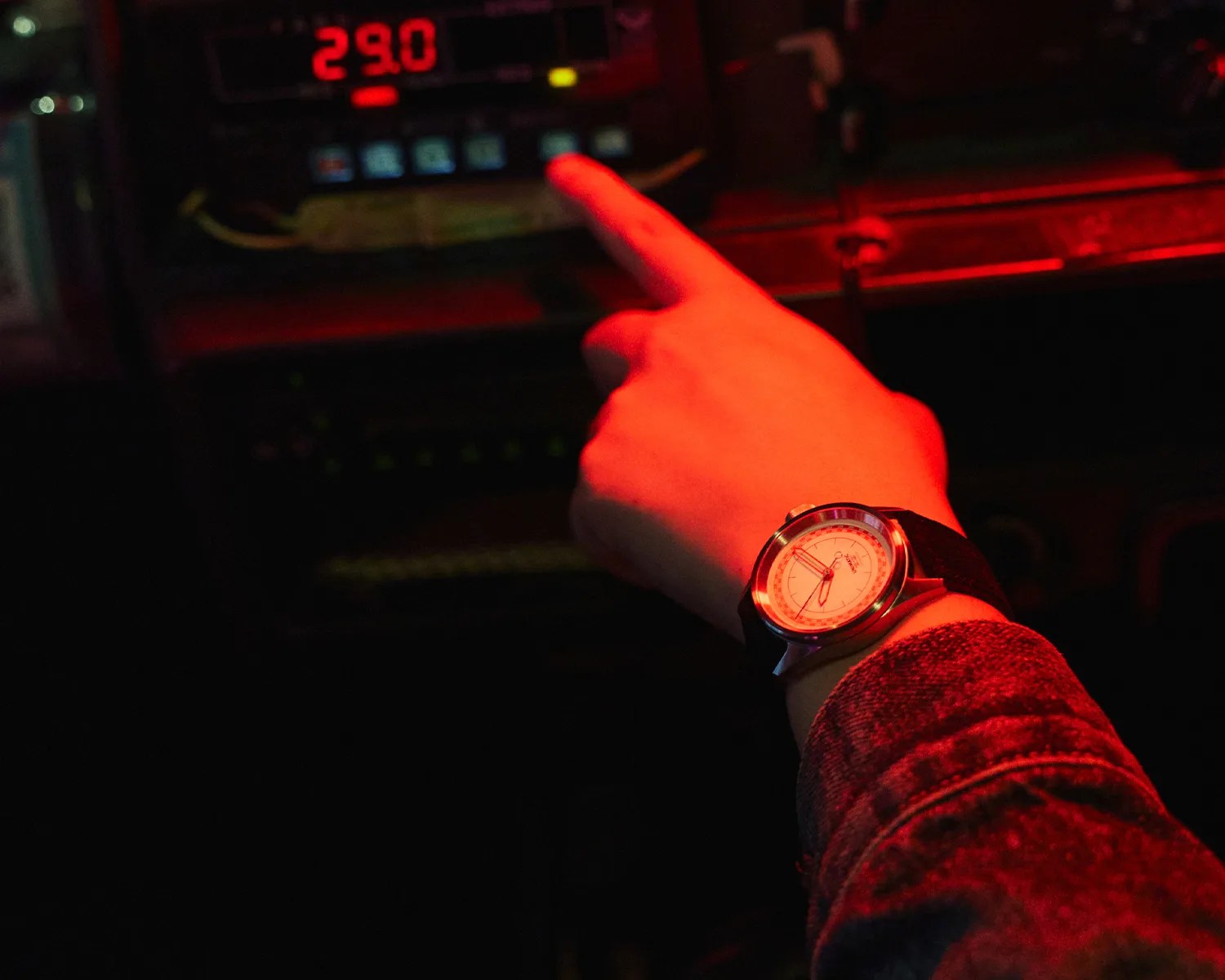 Hand wearing a wristwatch with a white dial and black strap pointing at a digital display showing 29.0 in red light.
